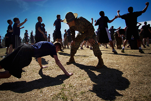 Une photographie montre une personne faisant des pompes tandis qu'un chef militaire se tient au-dessus d'elle ; d'autres personnes font des jumping jacks en arrière-plan.